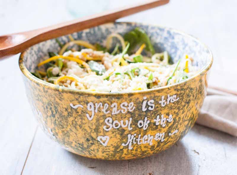 A blue, white, and yellow ceramic bowl with "grease is the soul of the kitchen" written on it in white glaze, with a summer squash and feta salad inside, with a light brown wooden spoon on top, on a white wood surface with a folded off-white cloth to the right of the frame.
