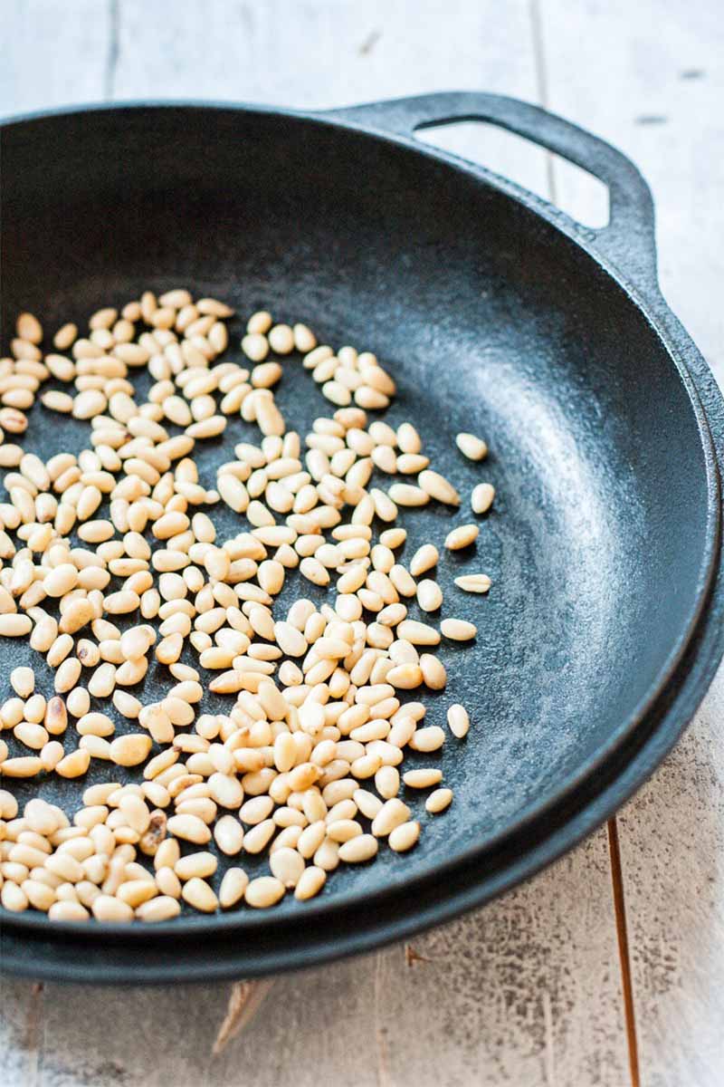 Toasted pine nuts in a large nonstick frying pan, on a white weathered wooden surface.