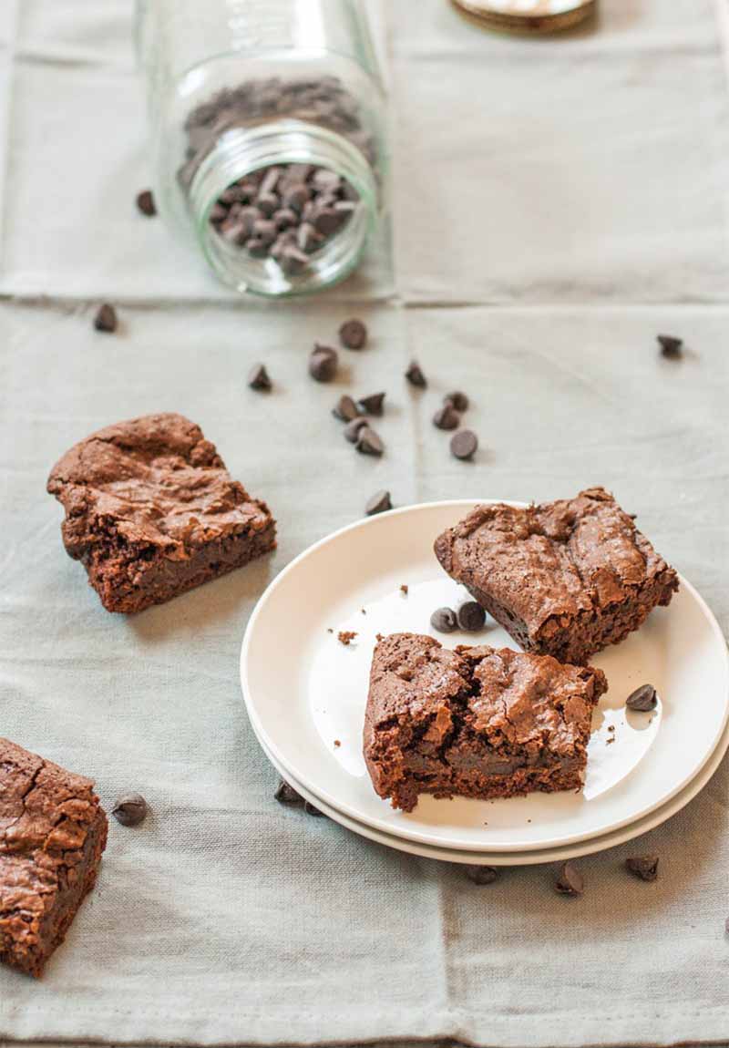 Two brownies on a stack of two white plates, and two more on a wrinkled gray linen tablecloth, with a tipped over mason jar of chocolate chips scattered across the table.