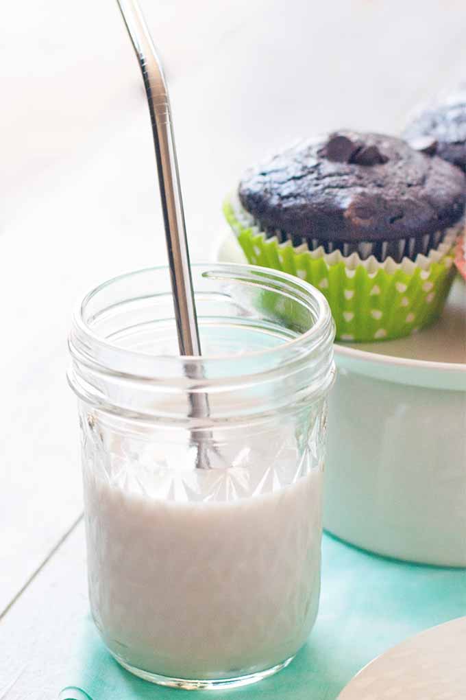 A mason jar of milk with a metal straw stuck into it for drinking is in the foreground, with a white pedestal serving platter of double chocolate muffins in polka dot green paper liners is in the background, on a white wood surface with a pale aqua cloth.