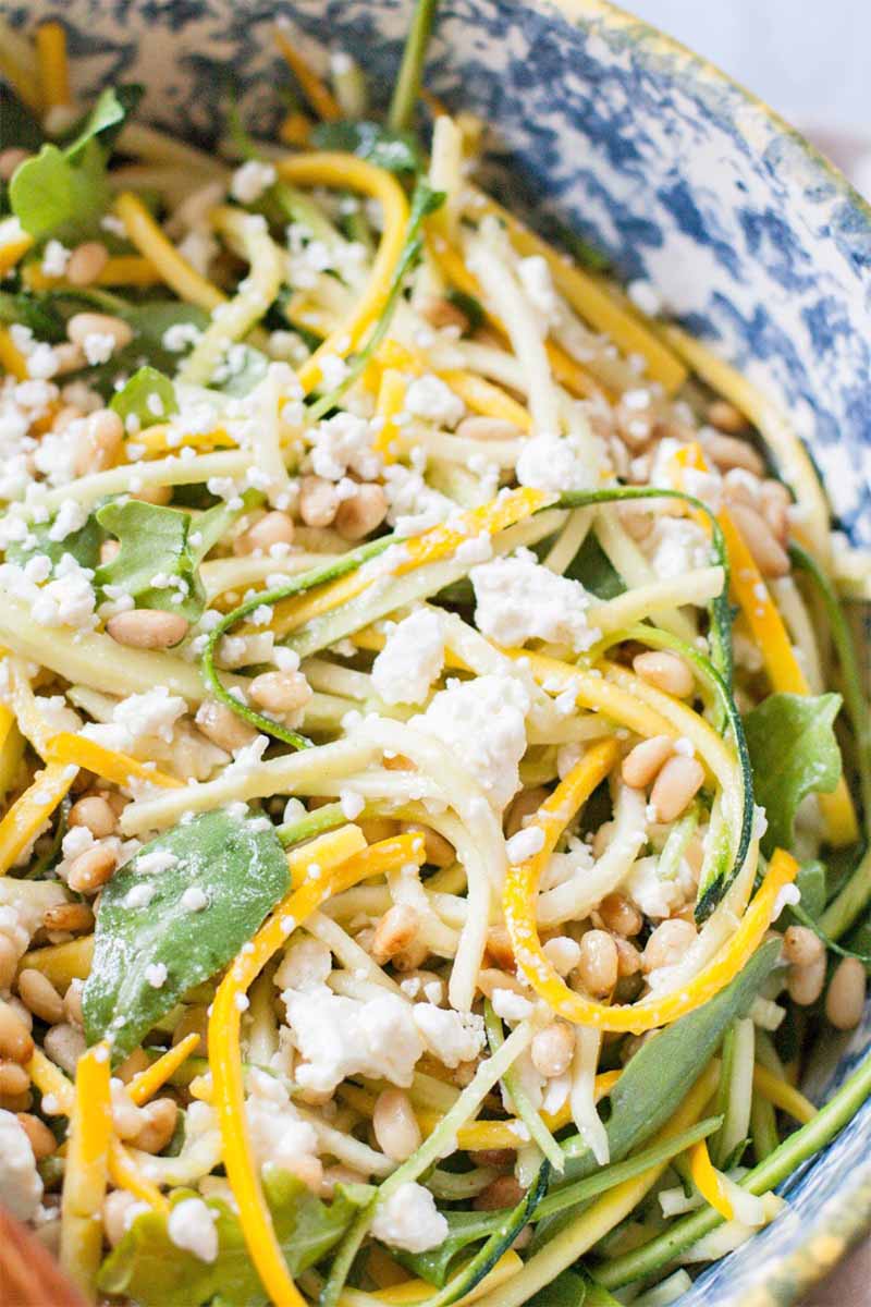 Closeup of a julienned summer squash and arugula salad, with crumbled feta and toasted pine nuts, in a blue and white ceramic bowl.
