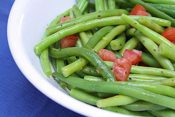 Closely cropped horizontal image of Thyme Seasoned Green Beans with Tomatoes in a white bowl, on a blue cloth surface.