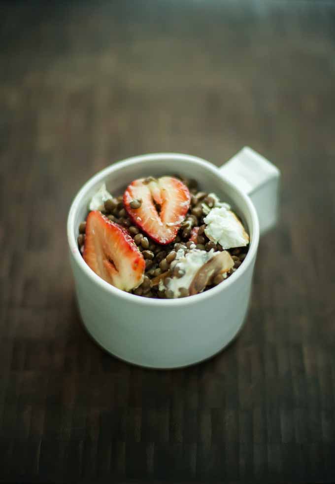 A white porcelain mug full of a recipe which includes Lentils with Shallots, Strawberries, and Goat Cheese. On a dark, wooden background.