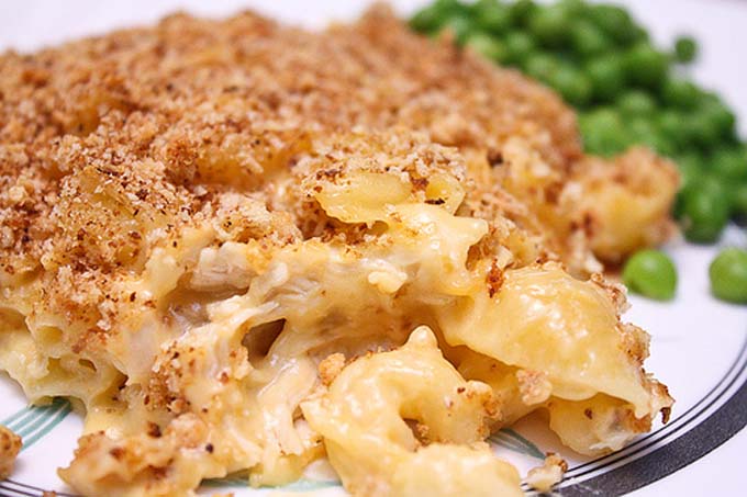 Horizontal image of chicken mac and cheese with a breadcrumb topping on a white plate next to peas.