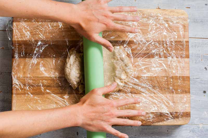A pair of hands rolls out the dough for the empanadas using a rolling pin and wooden cutting board. Top down view.