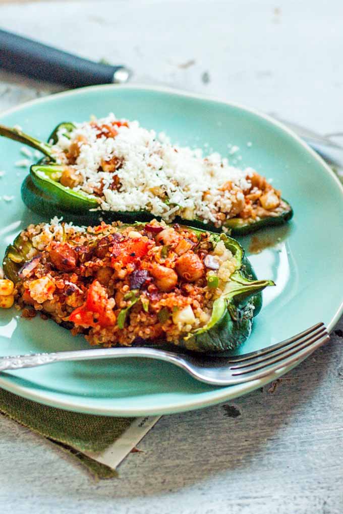 Two stuffed pasilla peppers on a blue-green plate with a fork, one of which is topped with grated cheese, on a white wood background in bright light.