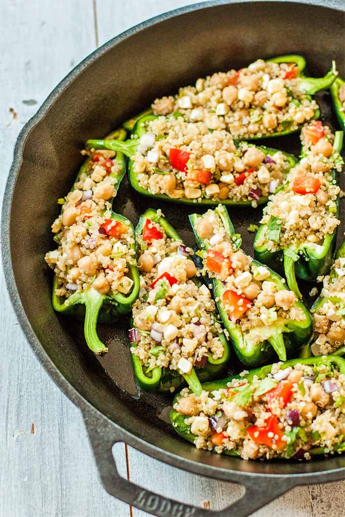 Nine halves of green poblano pepper filled with a quinoa and vegetable mixture and arranged cut-side up in a large black cast iron skillet, on a whitewashed wood background.