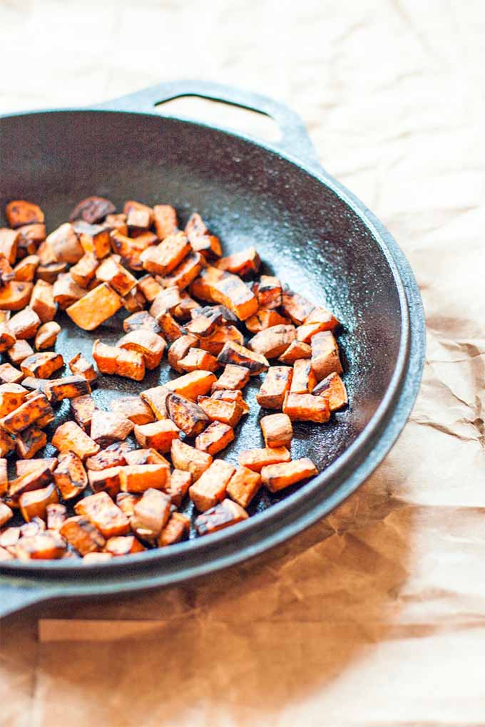 Vertical image of a cast iron pan of browned pieces of orange sweet potato, on a crumpled brown paper background.