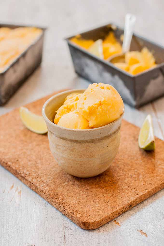 A cup of mango, lime, and cream cheese sherbet with two loaf pans full of the same in the background.