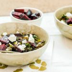 Three white ceramic bowls, two large and one small. The former are filled with kale, farro, beets, and goat cheese. The latter contains roasted beets. on a white tile surface with a green and white napkin with a floral pattern.