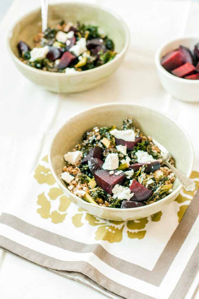 A white ceramic bowl of farro, kale, roasted beets, and crumbled goat cheese, with another bowl of the same with a spoon stuck into it in the background, beside a smaller white bowl of more beets, on a white tile surface with a green, white, and brown patterned paper napkin.