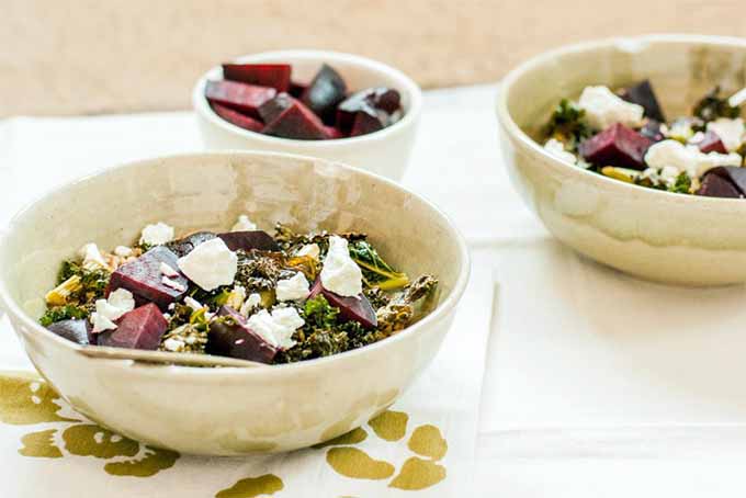 Three white ceramic bowls, two large and one small. The former are filled with kale, farro, beets, and goat cheese. The latter contains roasted beets. on a white tile surface with a green and white napkin with a floral pattern.