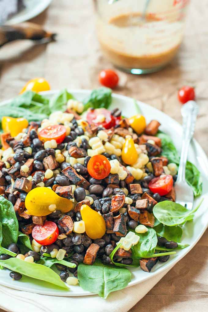 Closely cropped vertical image of a white dinner plate of spinach, black beans, grilled corn, roasted sweet potatoes, and small red and yellow tomatoes, with a small glass pitcher of miso dressing, whole red grape tomatoes, and a corn husk in the background, on a surface topped with a large piece of brown paper.
