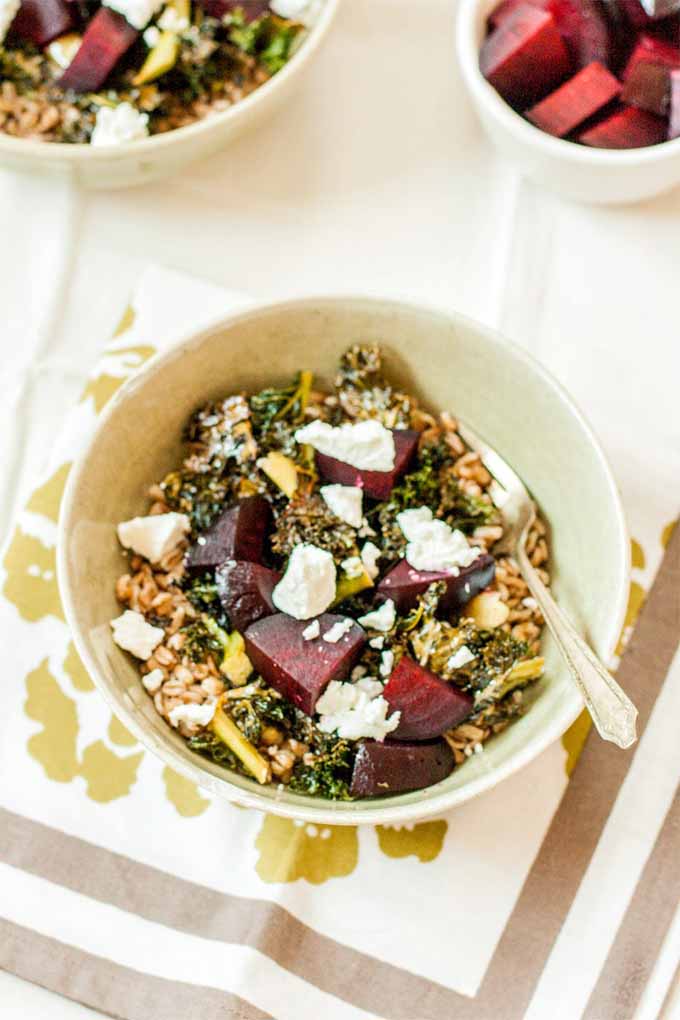 Top-down view of a white ceramic bowl of farro topped with roasted beets and kale, and crumbled feta cheese, with a silver spoon, on a green, white, and brown floral-patterned napkin, with another bowl of food and a bowl of just beets at the top of the frame.