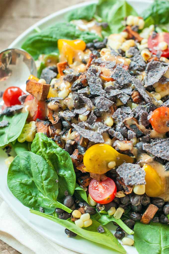Closeup of a spinach salad with red and yellow cherry tomatoes, roasted sweet potatoes, black beans, grilled corn, and crumbled blue corn chips, on a white plate on top of a folded white cloth napkin.