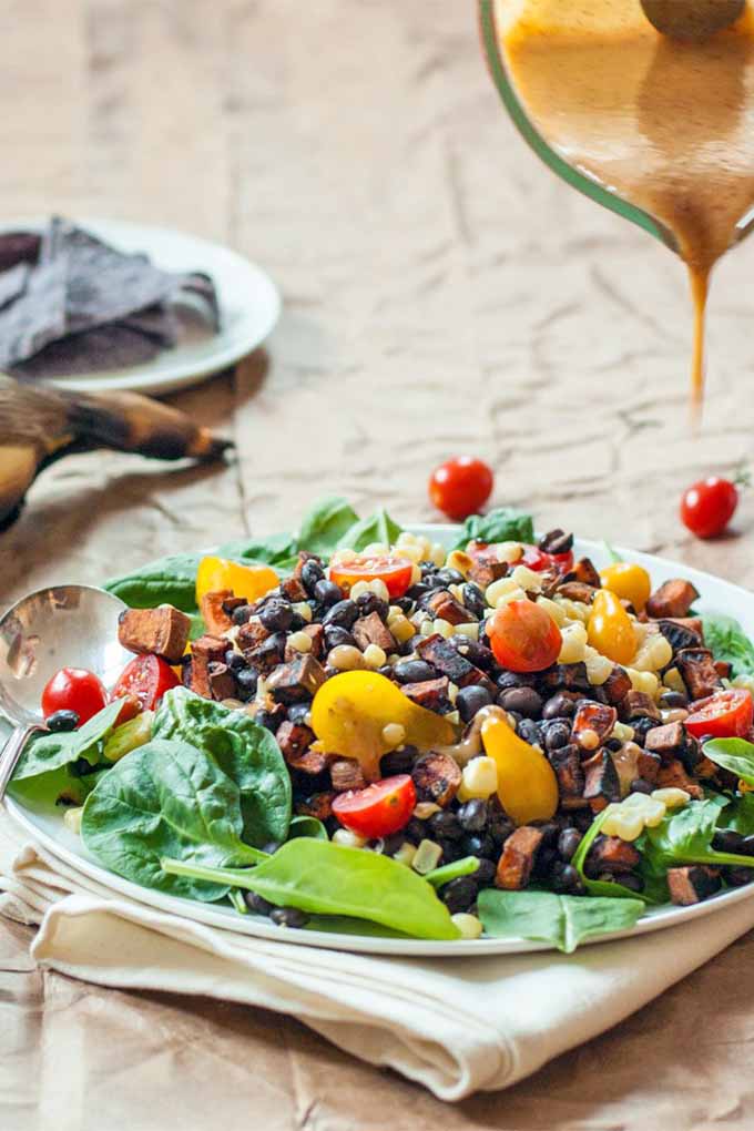 Vertical image of a bed of spinach on a plate on top of a folded white cloth napkin, topped with halved red and yellow cherry tomatoes, black beans, and roasted sweet potatoes, with a beige-colored miso dressing being poured from a pitcher at the top right of the frame, and scattered tomatoes, a plate of blue corn chips, and an ear of grilled corn in the background.