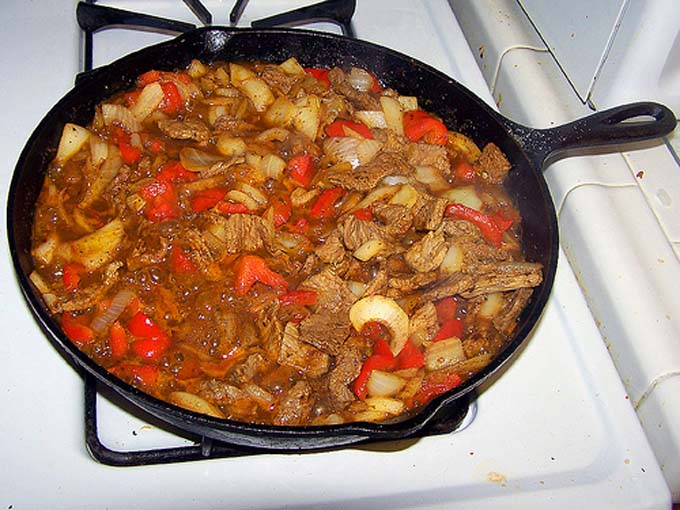 A beef stew with red bell peppers and onions cooks in a large cast iron pan on a gas stove, next to a white tile countertop.