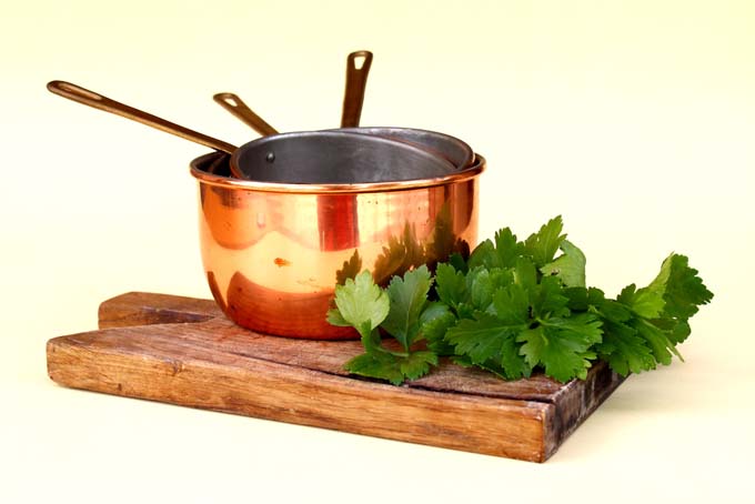 Copper cookware on a thick wooden cutting board with sprigs of flat leaf parsley, on a pale yellow background.