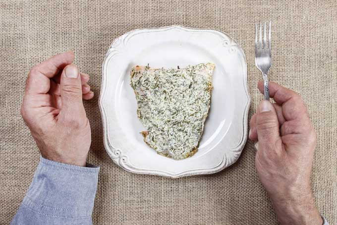 Horizontal image of hands, one holding a fork, next to a white plate with salmon covered in a creamy dill sauce