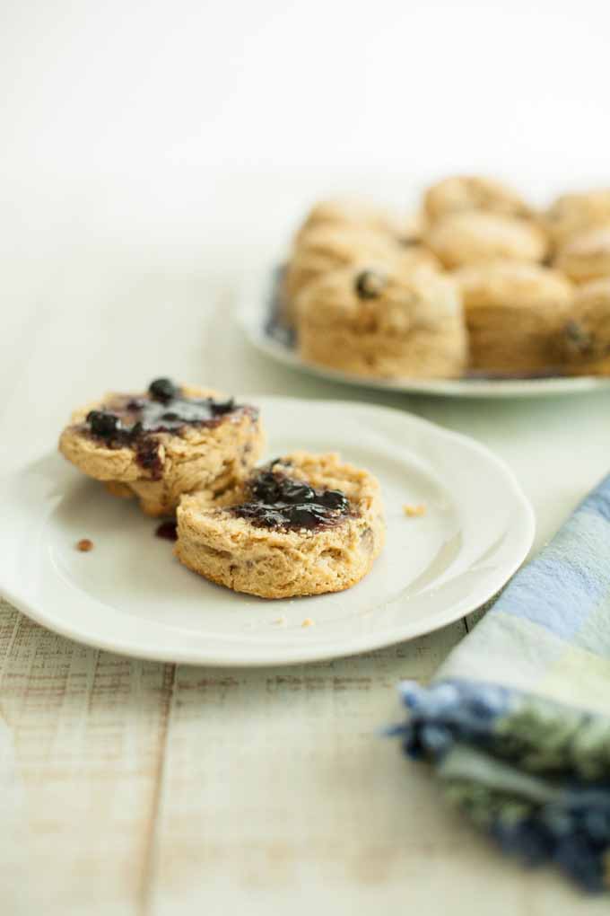 A single blueberry wholegrain kamut scone split in two with jam applied to both pieces. The remainder of the scones are on a plate in the diffused background.