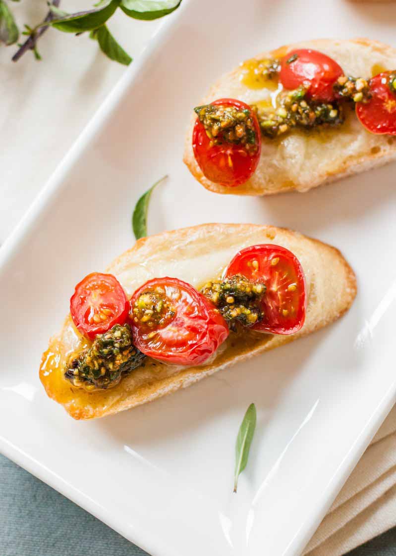 Top-down view of two slices of Italian bread topped with cheese, cherry tomatoes, and a pistachio-based pesto sauce on a square, white porcelain plate.