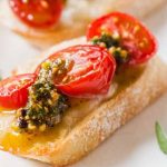 Macro view of a slice of Italian bread made into a bruchetta with Camembert cheese, a pistachio-based pesto sauce, and cherry tomatoes.