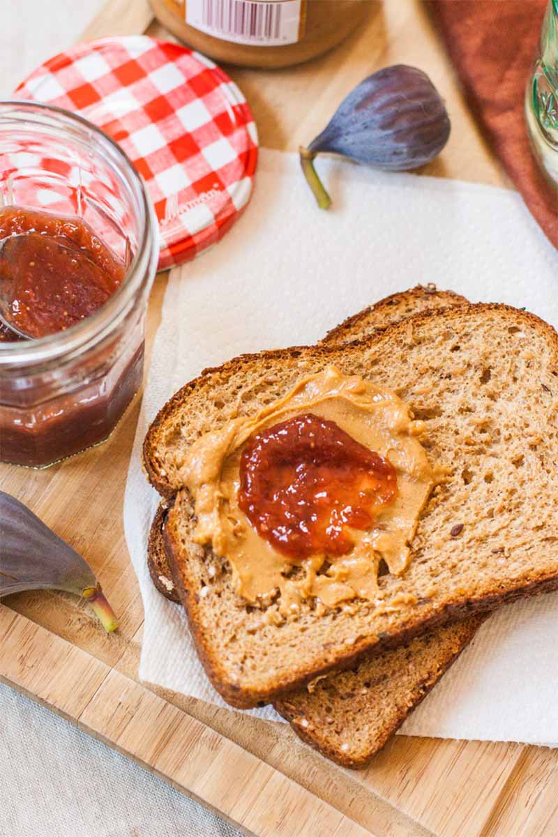 Vertical image of two pieces of brown bread with peanut butter and jelly on top, with a jar of jam and fresh figs on a wood cutting board.