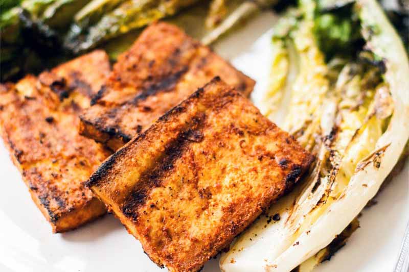Closeup of three rectangular pieces of marinated and grilled tofu beside wilted and charred romaine lettuce, on a white plate.
