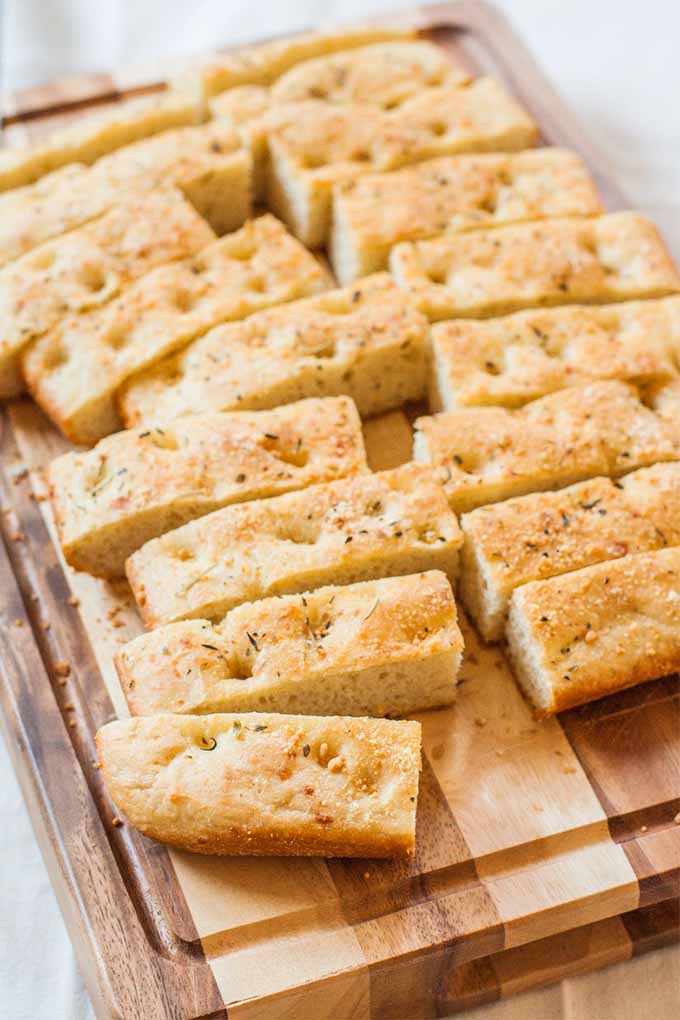 Golden brown focaccia bread topped with parmesan, sliced into narrow rectangles, on a wood cutting board.