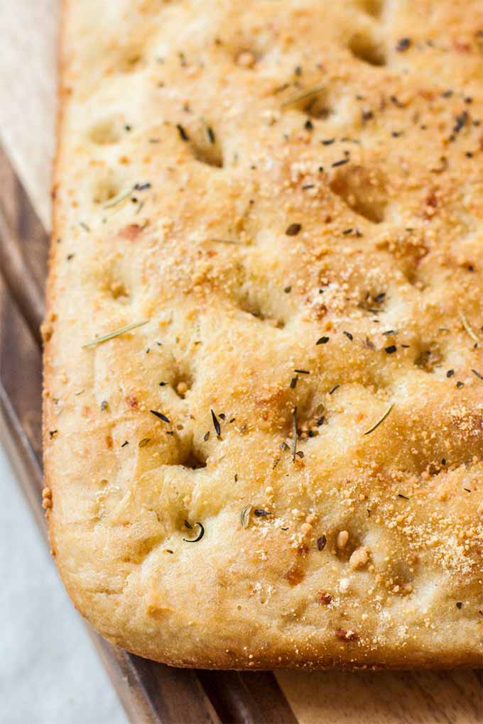 Closely cropped closeup of a rectangular, flat loaf of focaccia bread, with indentations in the top in a grid attangement, sprinkled with herbs and grated parmesan cheese, on a wood cutting board.