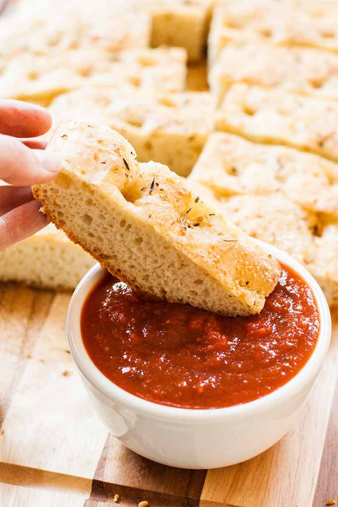 Closely cropped image of fingers holding a narrow rectangular slice of focaccia, dipping it into a white ceramic ramekin of red marinara sauce, on a wooden cuttinb board topped with more slices of the bread.