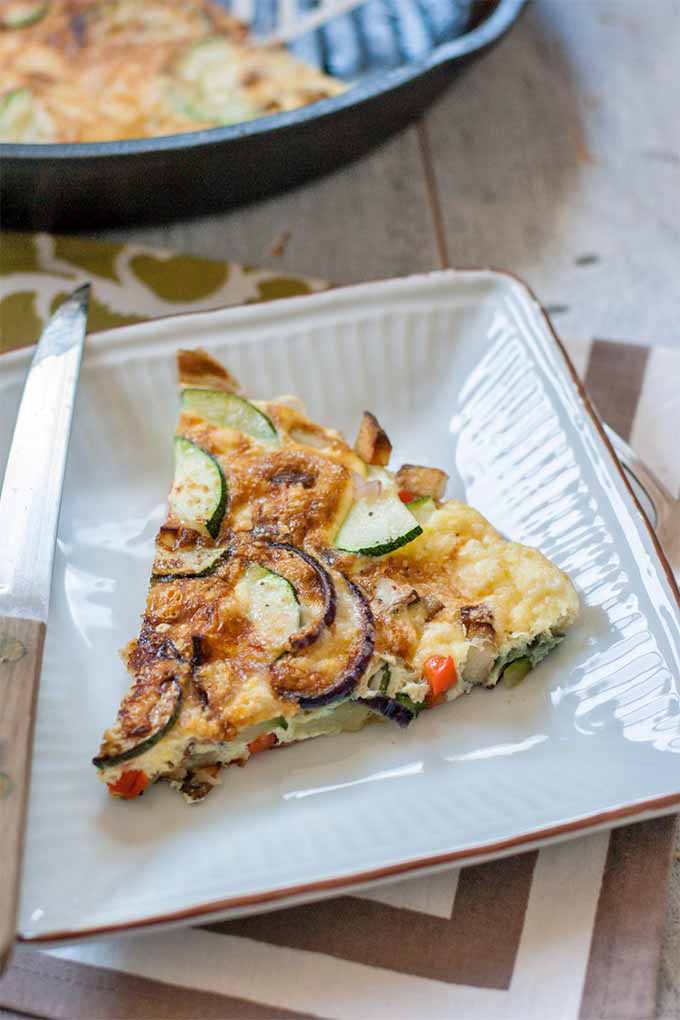 A triangular slice of vegetable frittata on a square white plate with a wood-handled serrated steak knife, on a patterned place mat with a cast iron pan of the remainder of the egg dish in the background, on a white stained wood table.