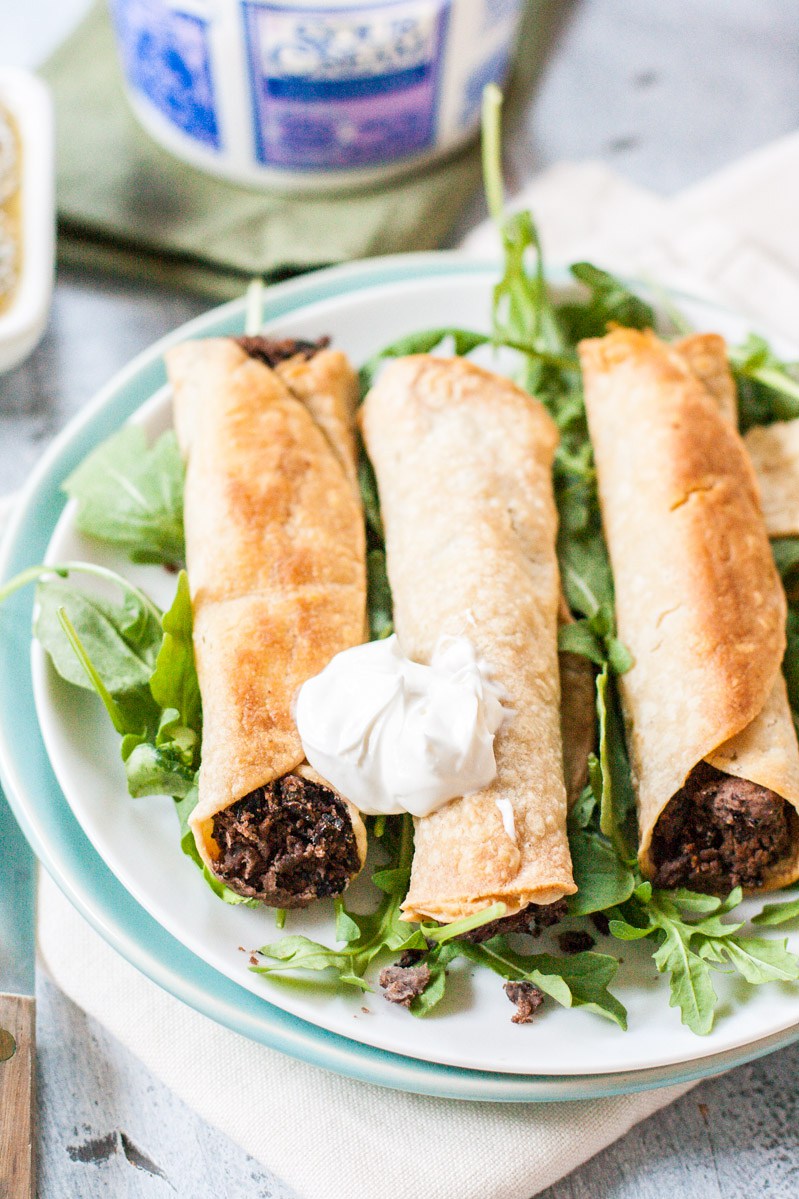 Three vegan and gluten-free slow cooked baked taquitos made with slow cooked black beans and charred poblano peppers. A dollop of vegan sour cream has been added to the top.