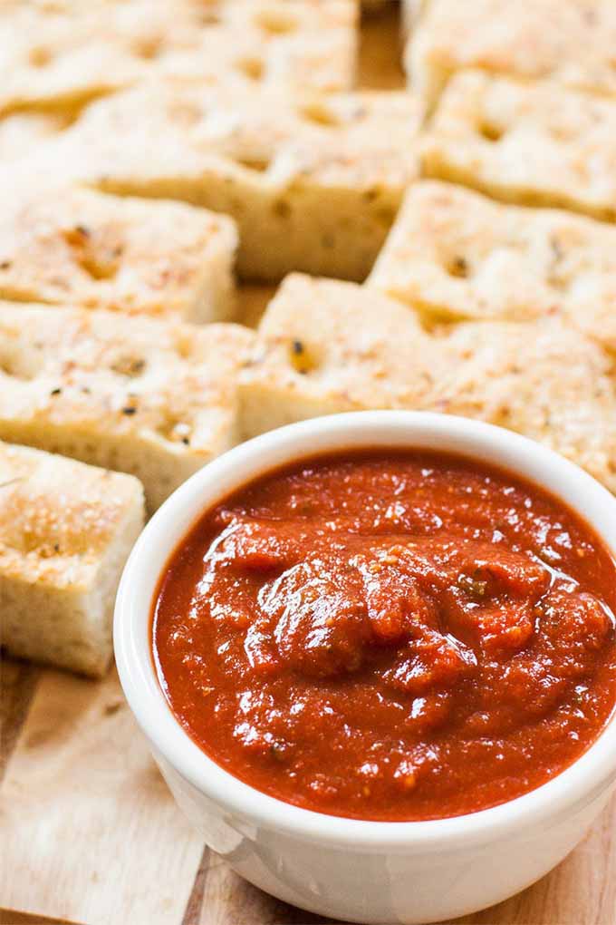 Slices of homemade focaccia bread on a wood cutting board with a white ceramic ramekin of red marinara sauce.