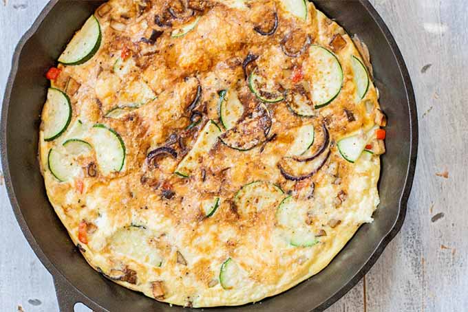 Top-down shot of a potato and zucchini egg frittata in a cast iron pan, golden brown on top, on a distressed wood background.