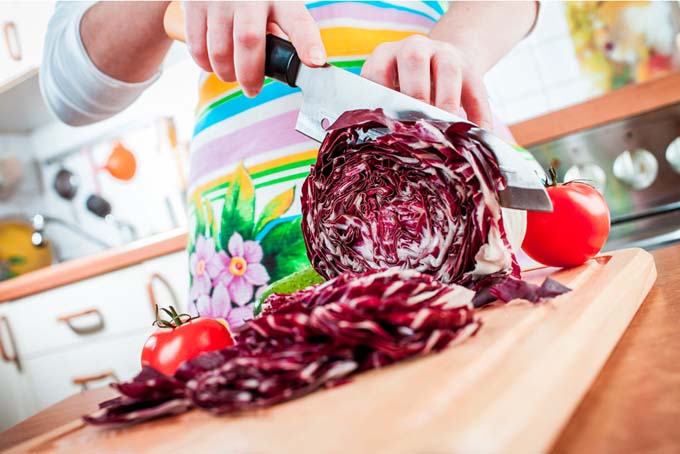 Woman's hand and chef's knife chopping up purple cabbage on wooden chopping board.