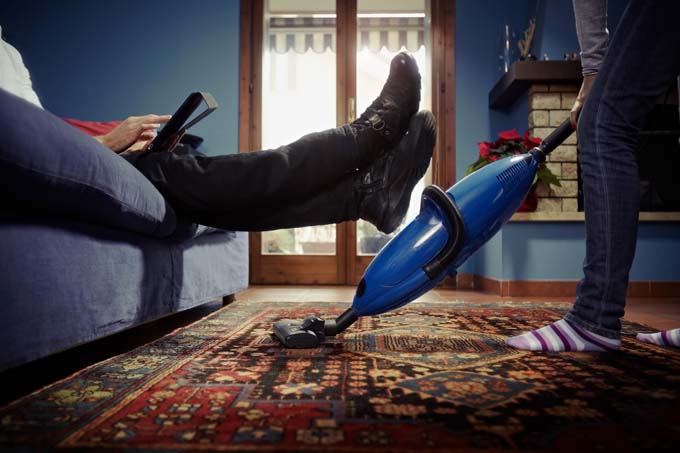 Man relaxing on couch while woman vacuums carpet