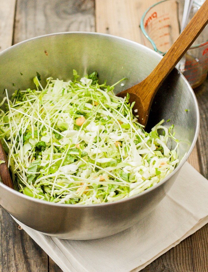 Oblique view of a stainless steel mixing bowl full of a cabbage and pea sprout salad. A wooden spoon is stuck inside of the bowl with the handle leading to the upper right.
