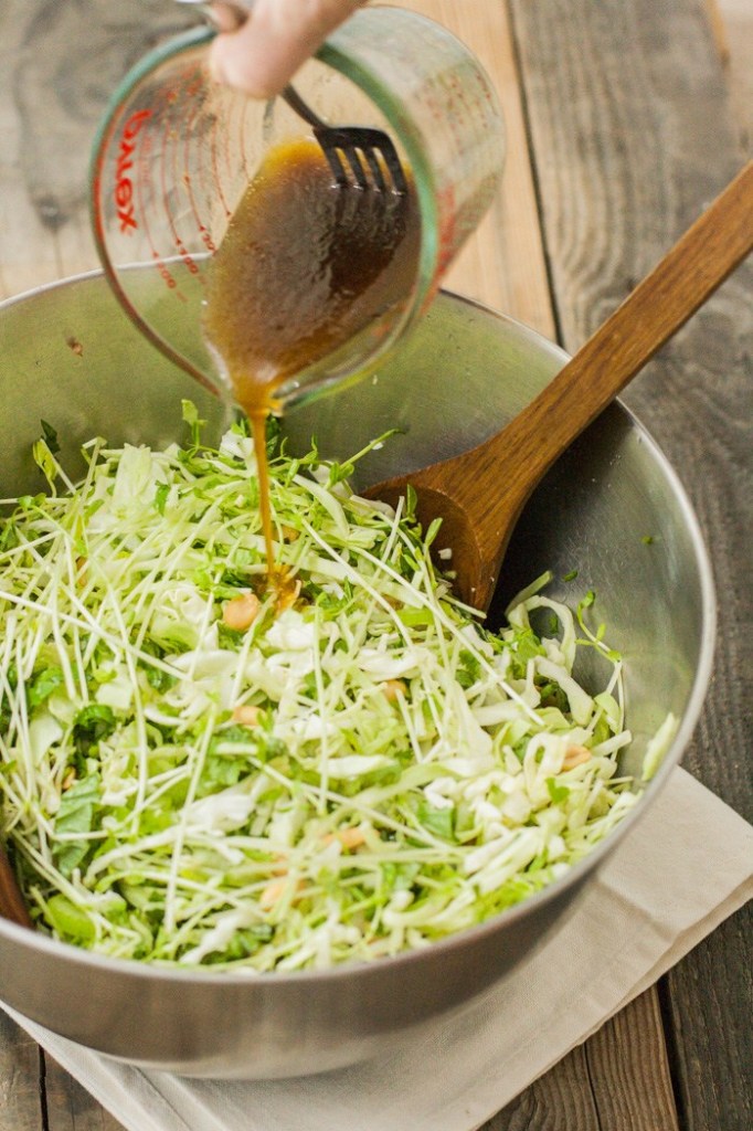 The dressing is being poured from a glass measuring cup over the salad in a large mixing bowl.