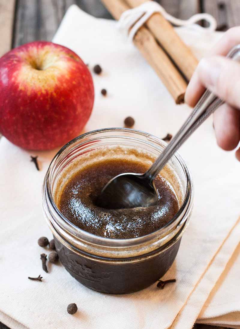 A close up of small 1/2 pint mason jar full of caramel sauce with a whole apple and cinnamon sticks placed in the background.