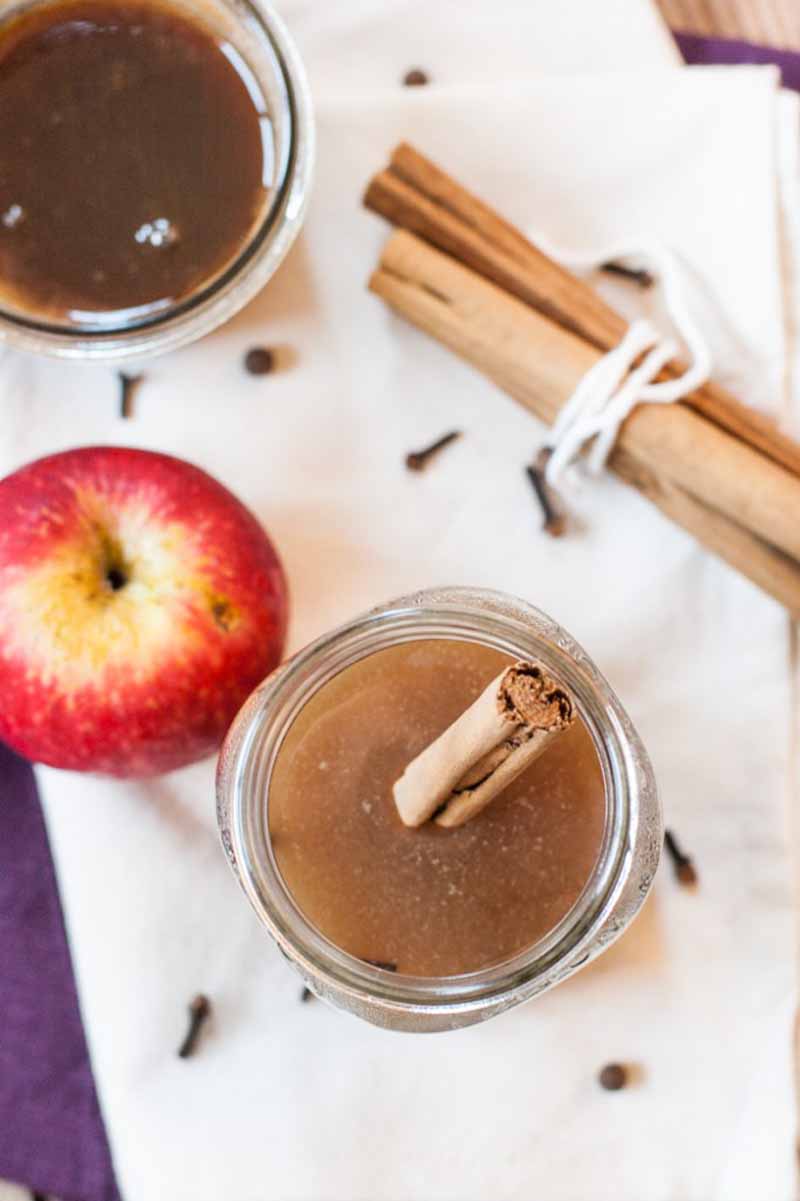 Top down view of an caramel apple cider drink, a jar of caramel sauce, a fresh apple, sticks of cinnamon, and cloves scattered about.