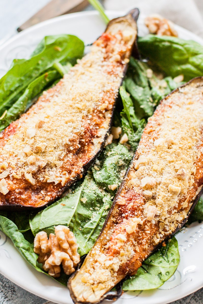 Closeup top-down view of two eggplant halves laying on a bed of spinach on a white porcelain plate. The halves have been stuffed with marinara and topped with a vegan-friendly Parmesan replacement that is made of nutritional yeast and walnuts.