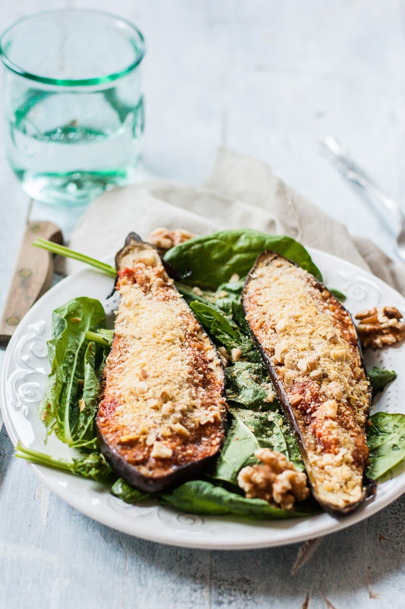 Oblique view of two eggplant halves stuffed with marinara sauce and topped with a vegan Parmesan cheese. The halves are laying on a bed of greens on a white ceramic plate.