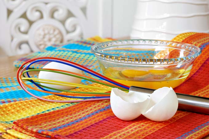 Eggs cracked into a bow with a wisk next to them. Shells in front. On colorful rainbow colored woven mat.