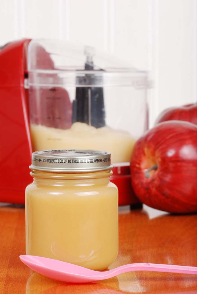 A babby food jar full of apple sauce in the foreground with food processor and apples in the background