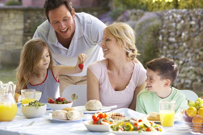arrtactive couple with son and daughter in an outside brunch consiting of healty foods; daughter is holding strawberry in the air and parents are laughing