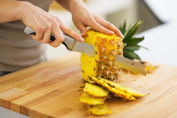 Humand hands and knife peeling the skin off a pinapple on top a cutting board as part of preparation proceess