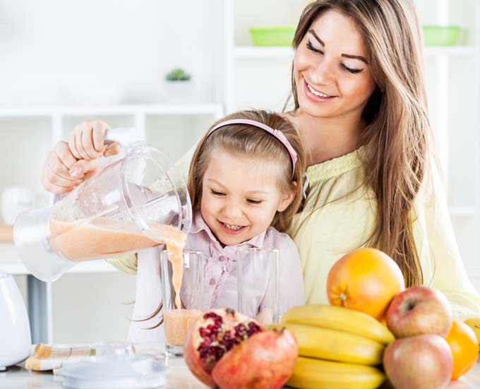 Mother and daughter pour a smoothie from a blender; they are behind a kitchen counter that includes oranges, kiwi,bananas and other fruit on its surface