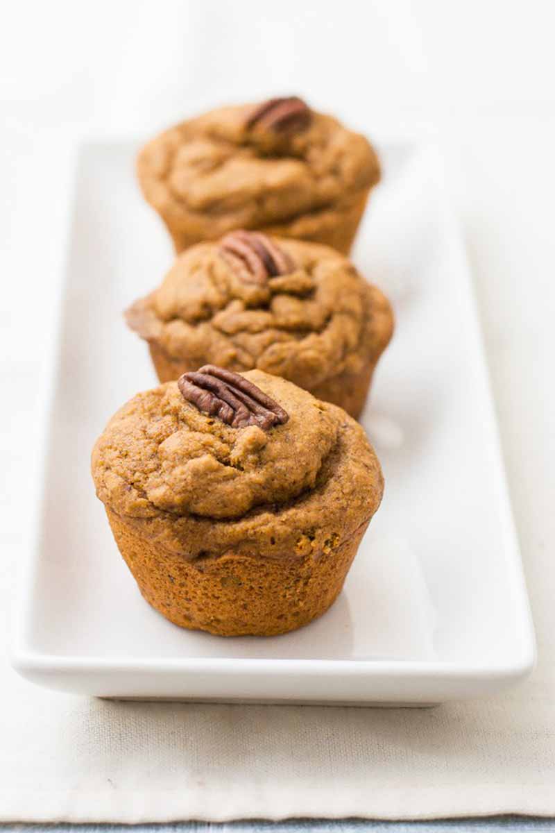 Vertical image of three pumpkin muffins with pecans on top of a white platter.