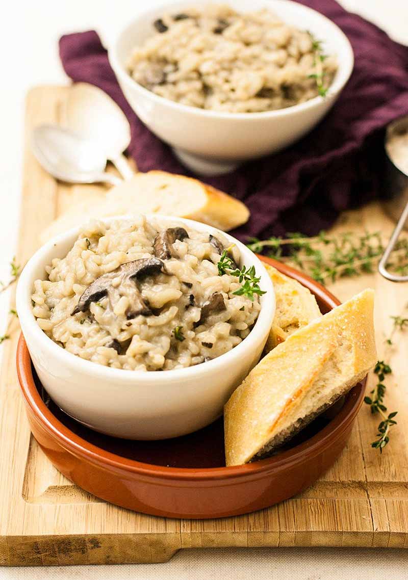 Two white ceramic bowls full of a vegan mushroom and herb rice dish with crusty Italian bread on the side.